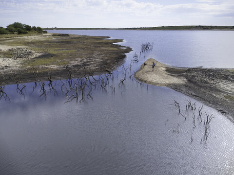 Aerial view of Colliford Lake Reservoir with a person standing on a sandbar surrounded by dead trees protruding from the calm water in Bodmin, Cornwall, United Kingdom.
