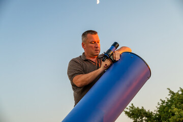 a man adjusting a large blue telescope outdoors during twilight with a crescent moon visible in the sky. © Andrey