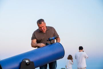 a man adjusting a large blue telescope outdoors against a clear twilight sky, with other people standing in the background. © Andrey