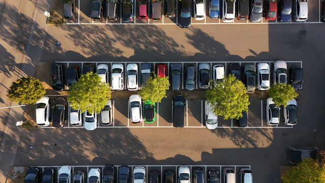 Aerial view of a parking lot with numerous cars parked in marked spaces, surrounded by green trees casting shadows on the asphalt in Schaffhausen, Schaffhausen, Switzerland.