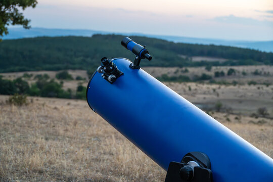 A blue reflecting telescope on a Dobson mount is set up in a dry field overlooking a rolling landscape at sunset.