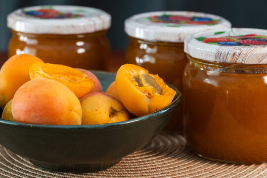 A black ceramic bowl filled with fresh whole and halved apricots sits in front of three glass jars of homemade apricot jam on a woven mat.