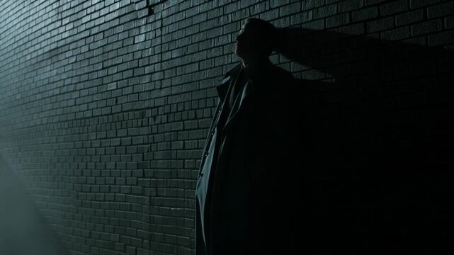Man in dark coat leans against brick wall in shadow under low side lighting