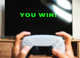 Blurred gamepad in hands against TV screen with green sign You Win © Igor Barilo