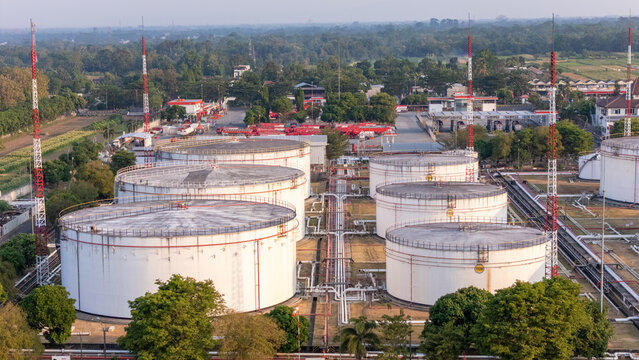 Aerial view of large white cylindrical fuel storage tanks and industrial pipelines at a facility surrounded by lush green trees in Indonesia.