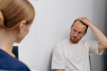 Male patient consulting doctor about hair loss while touching scalp in clinic for consultation...