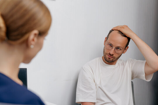 Male patient consulting doctor about hair loss while touching scalp in clinic for consultation about medical transplant