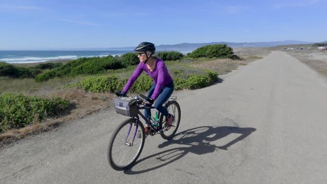A senior woman bikes along a paved trail next to the ocean in Fort Bragg, California. She enjoys the outdoor adventure under a clear sky.