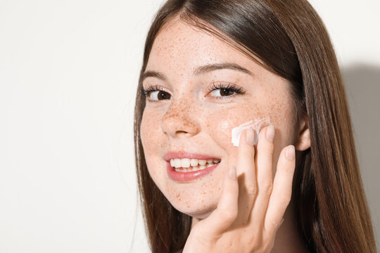 Beautiful teenage girl with freckles applying cream onto her face against white background, closeup. Space for text