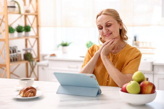 Smiling senior woman looking at tablet during coffee break at white marble table in kitchen