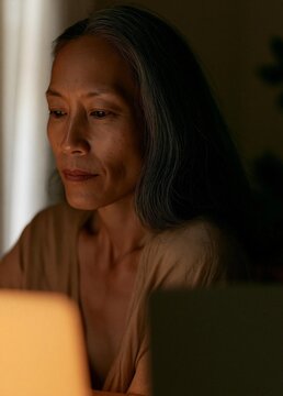 Portrait of a middle-aged asian woman sitting in front of a laptop. she is looking off to the side with a thoughtful expression on her face.