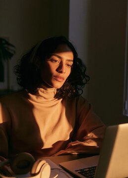 Young woman sitting at a desk with a laptop in front of her. she is wearing a beige turtleneck sweater and has a pair of headphones resting on her lap.