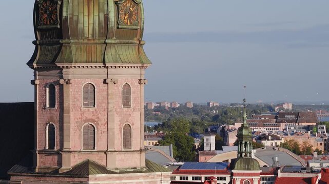 Aerial view shows the historic clock tower of Saint Peter church, city rooftops, and the Daugava River under clear daylight in Riga, Latvia.