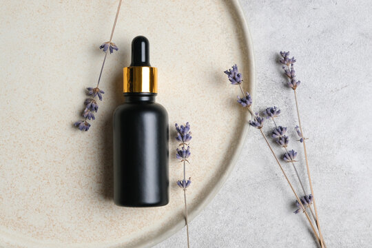 Black glass dropper bottle with golden cap placed on a beige plate surrounded by dried lavender flowers on a textured gray surface