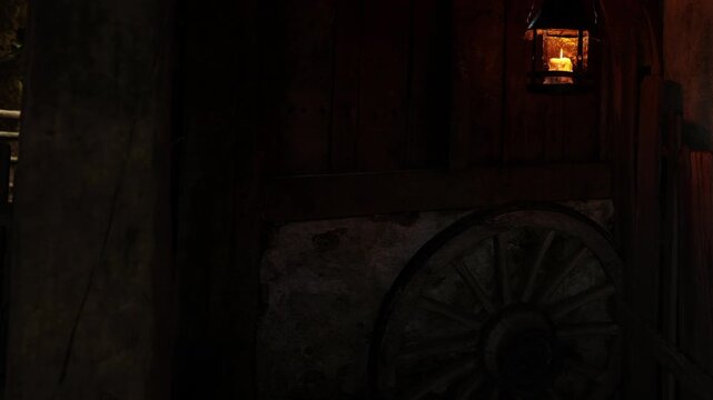 A room shows a wooden wagon wheel placed against a wall. A hanging lantern provides soft light in the dark space, creating a rustic atmosphere at night.