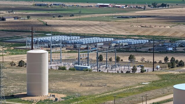 Aerial view of an electrical substation and battery energy storage facility with rows of white containers and power lines set in a rural landscape in Frederick, Colorado, United States.