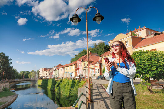 Young woman with red hair and sunglasses happily using her smartphone for navigation, enjoying a sunny city trip by a canal in Knjazevac, Serbia