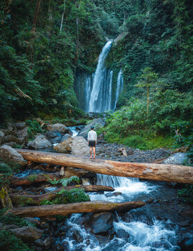 Aerial view of Tiu Kelep Waterfall cascading into a rocky stream with a person standing on a large fallen log amidst lush tropical forest in Bayan, West Nusa Tenggara, Indonesia.