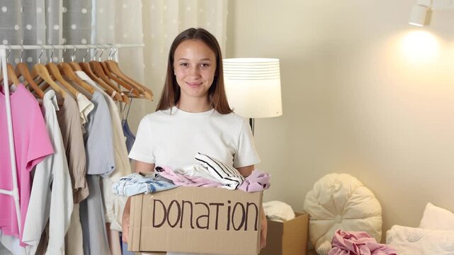 Teenage girl collecting and sorting her clothes for donation, placing garments into a cardboard box marked Donation.
