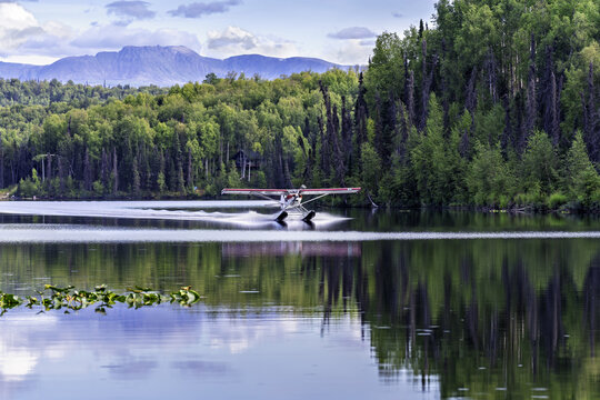 Aerial view of a seaplane landing on a calm lake surrounded by dense forest and distant mountains in Alaska, United States.
