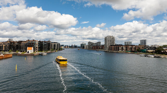 Aerial view of a yellow canal boat cruising through a waterway surrounded by modern apartment buildings under a blue sky with white clouds in Copenhagen, Denmark.