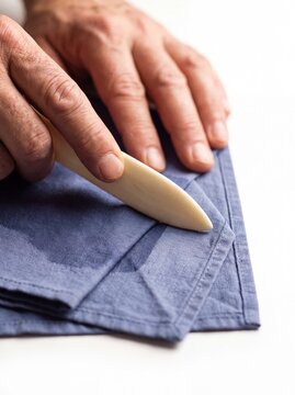 Artisan hands using bone folder tool on blue fabric, bookbinding craft process, traditional textile folding technique