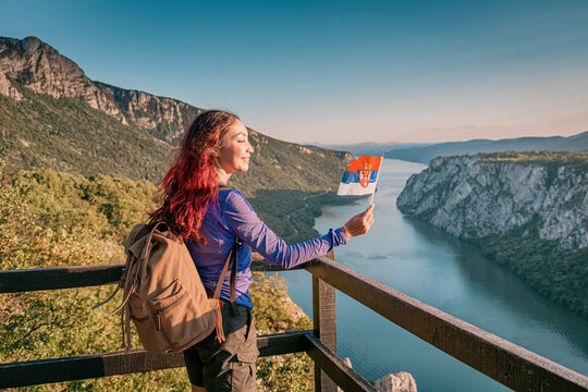 Joyful female hiker holds a Serbian flag at a dramatic Danube river, Iron Gates gorge overlook, radiating national pride and wanderlust. Travel in Djerdap park concept.