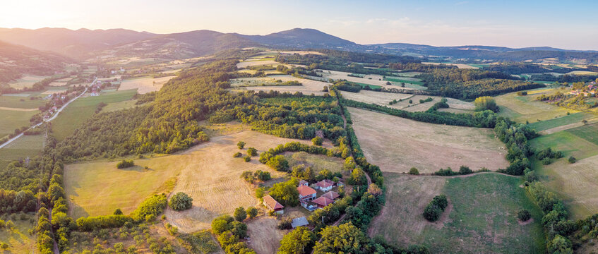 Aerial panoramic view of rolling countryside farmland with red-roofed homestead at golden sunset. Rural escape concept.