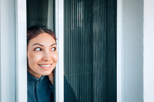 Smiling woman looking through a mosquito screen door onto a sunny balcony, enjoying fresh air and protection from insects during summer