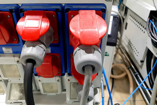 Industrial three phase power plugs and sockets on factory wall. Electrical cables connected to heavy duty red high voltage outlets in workshop background.