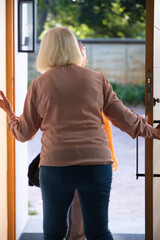 Naklejka premium Senior woman in pink blouse holding door in doorway with metal latch greeting woman carrying bag