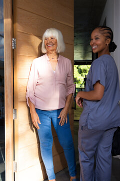 Senior female resident and adult female caregiver standing in home entryway, gray scrubs sunlight