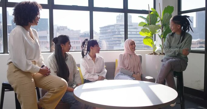 Female coworkers sharing ideas by windows, white-blouse woman leaning gesturing, drawing laughs
