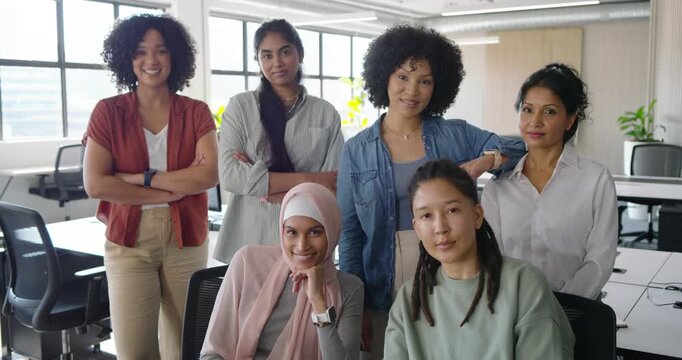 Diverse coworkers posing in open office by windows after camera cue, headscarf adjusting, smiling