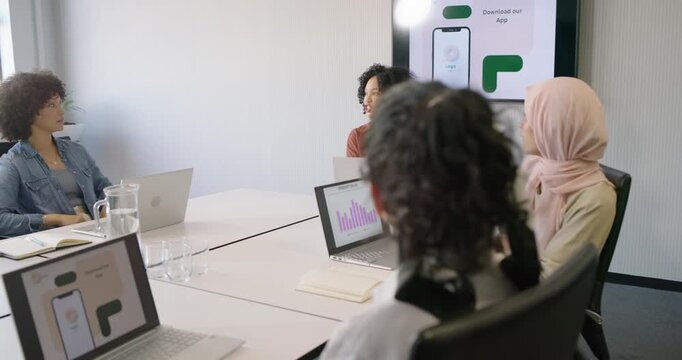 Diverse women reviewing app, presenter in rust cardigan pointing to wall mockup, others on laptops