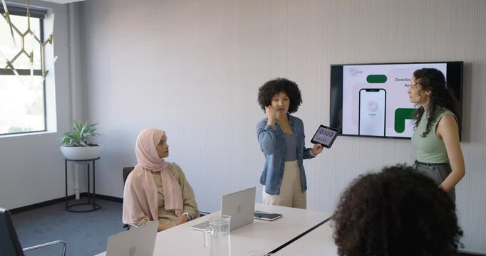 Diverse female team watching presenter rotating tablet showing app UI for project update in room