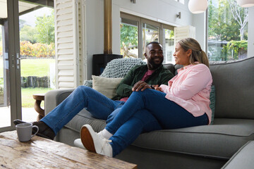 Naklejka premium Diverse couple sitting on grey sofa in living room holding hands beside coffee table with mug