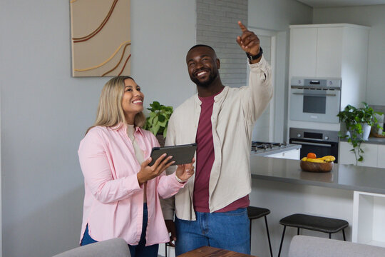 African American couple smiling, holding tablet and pointing upward in modern kitchen by island