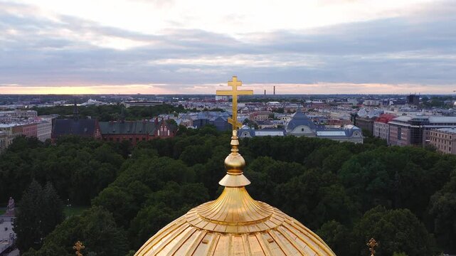 An aerial panorama captures the golden dome and cross of Nativity of Christ Cathedral with the Riga cityscape at sunset.
