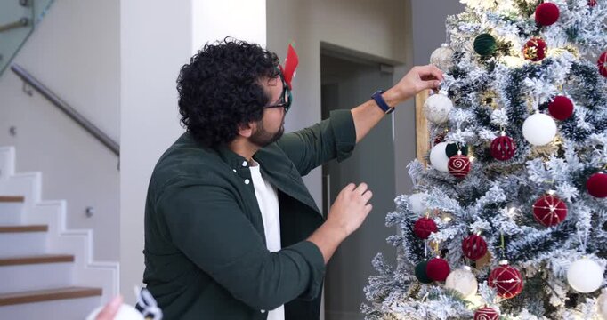 African American man reaching, decorating tree with bauble by stairs donning Merry glasses smiling