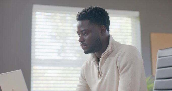 African American man in sweater at desk by blinds holding mug reading laptop then typing replies