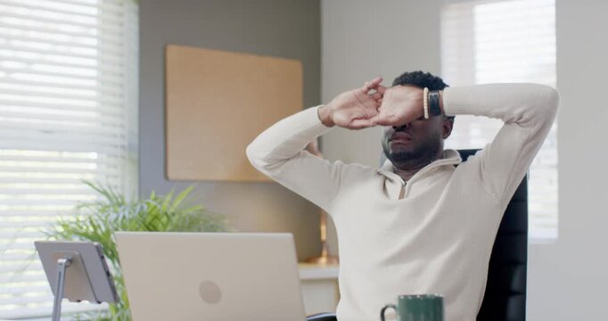 Man in cream pullover working at home office desk, focusing on laptop, stretching wrists for relief