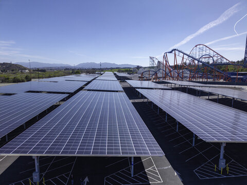 Aerial view of a large-scale solar panel array covering a parking lot with bifacial photovoltaic modules and a roller coaster in the background in Valencia, California, United States.