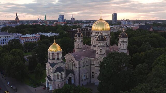 Aerial view of the golden domes of the Nativity of Christ Cathedral during sunset, surrounded by lush trees and the historic cityscape of Riga, Latvia.