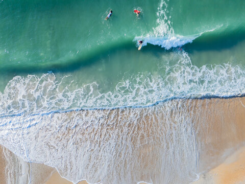 Aerial view of surfers riding waves in the turquoise ocean with white foam breaking on the sandy shore at Wrightsville Beach, North Carolina, United States.