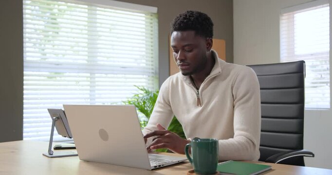 African American man typing on laptop at desk, pausing and stretching to relieve work tension