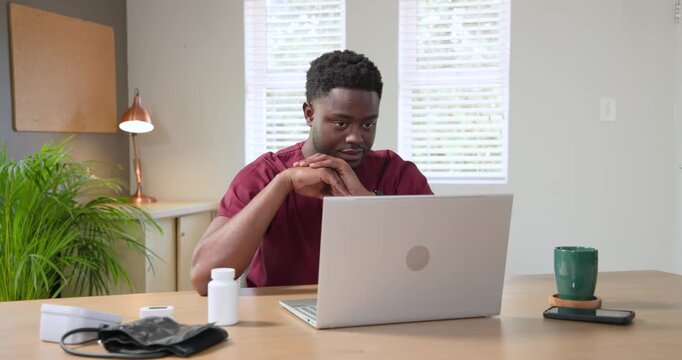 African American man in scrubs joining call at desk showing pill bottle to silver laptop