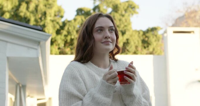 Adult woman standing on patio in white sweater holding glass cup, shifting gaze