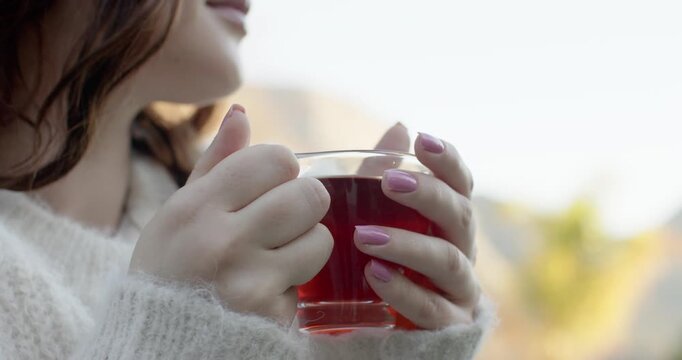 Breathing softly, woman is steadying glass cup of red tea on balcony, is warming hands
