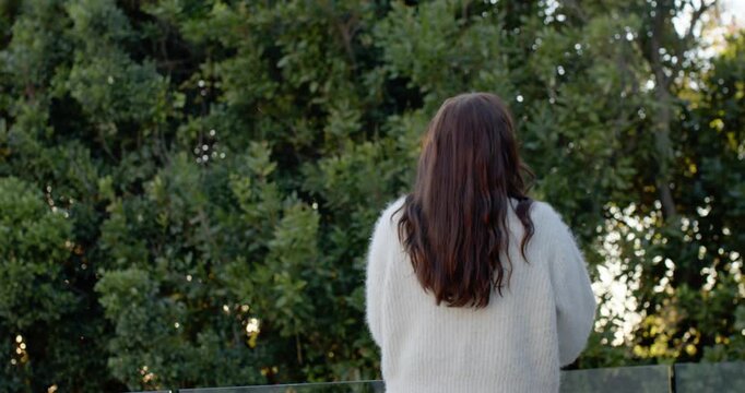 Light breeze is moving hair as woman is standing at glass railing watching sunlight through pines